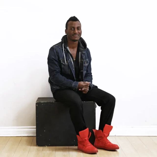 Young man sitting on box in front of white wall wearing red shoes and smiling at camera