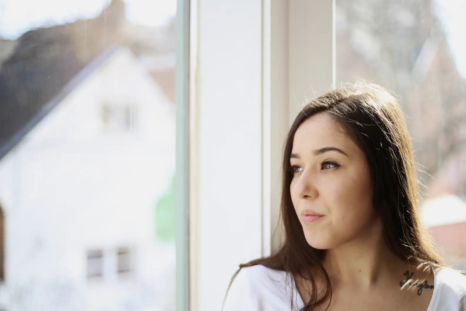 Young woman with long brown hair in white shirt looking out of sunny window