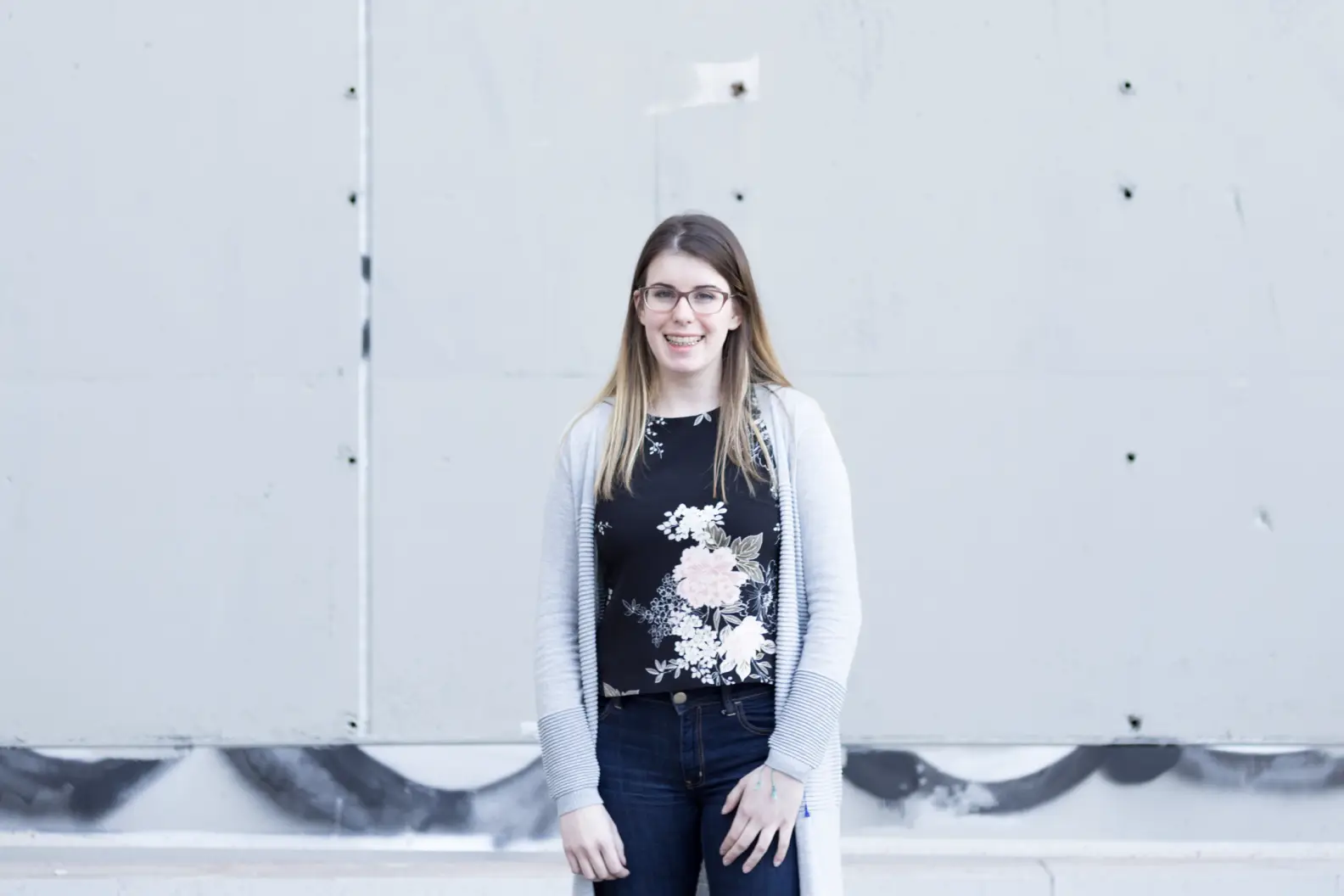 Young woman with long brown hair and glasses standing in front of grey concrete wall