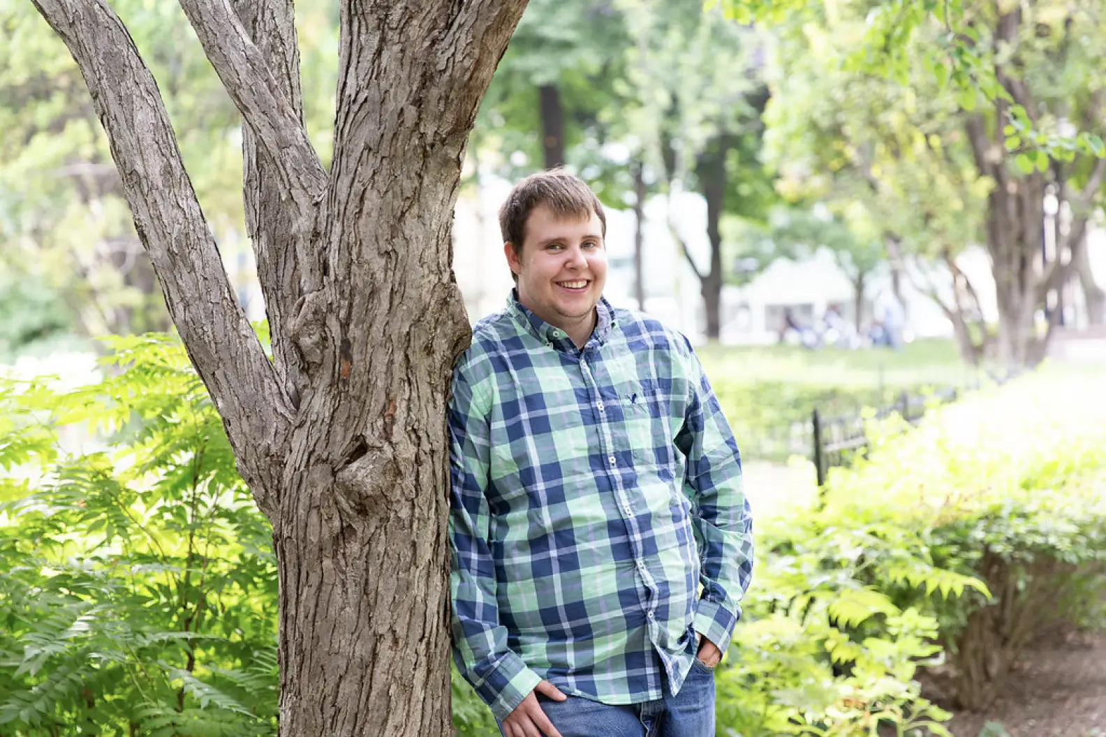 Young man in plaid shirt leaning against tree and smiling