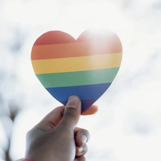 A hand holding a cut-out of a heart with a rainbow on it up to the sun