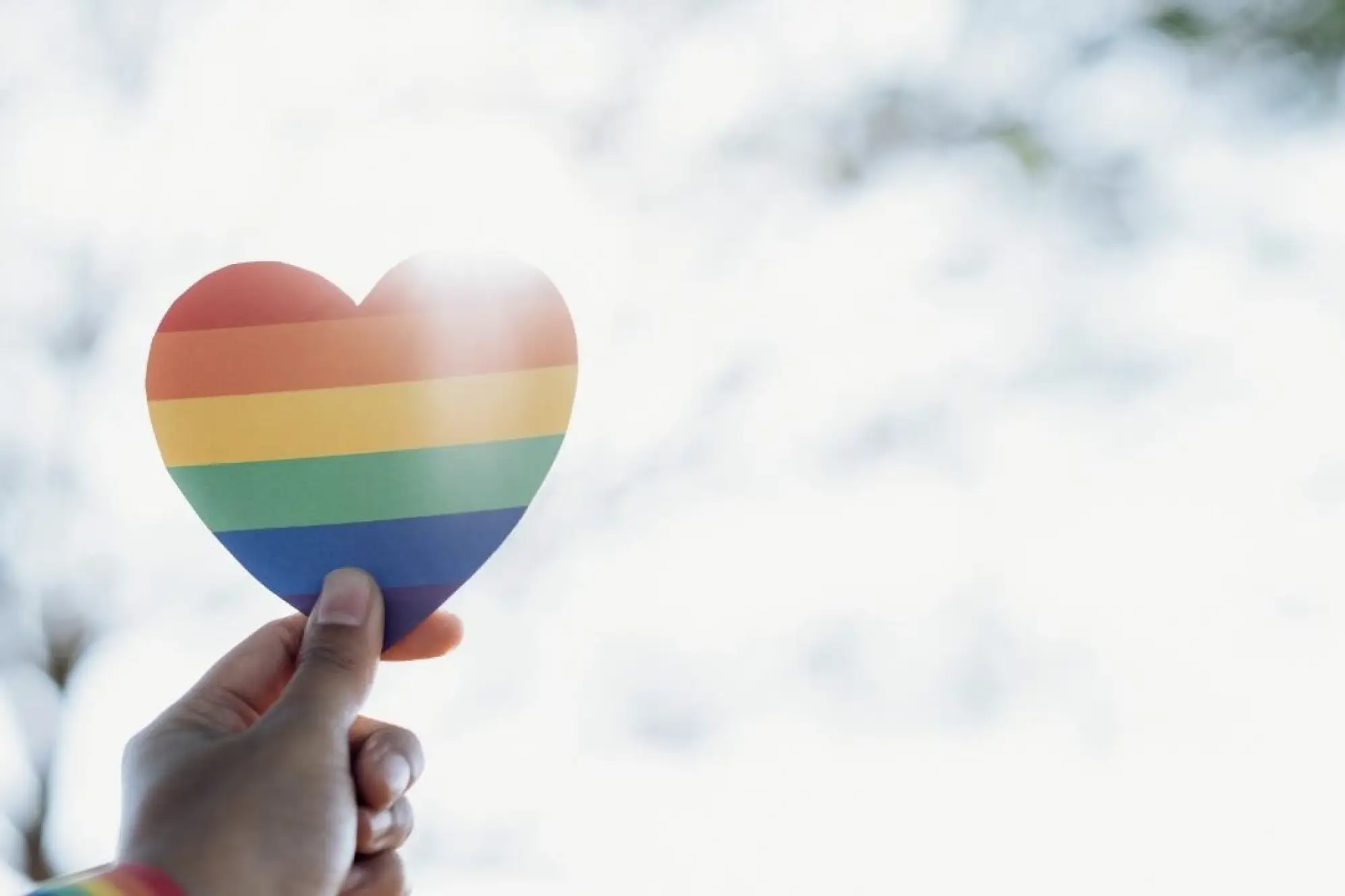 A hand holding a cut-out of a heart with a rainbow on it up to the sun