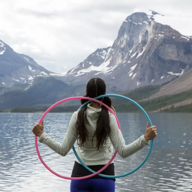 Person with two long pigtails and a blue and pink hoola-hoop standing in front of a lake with mountains in the background