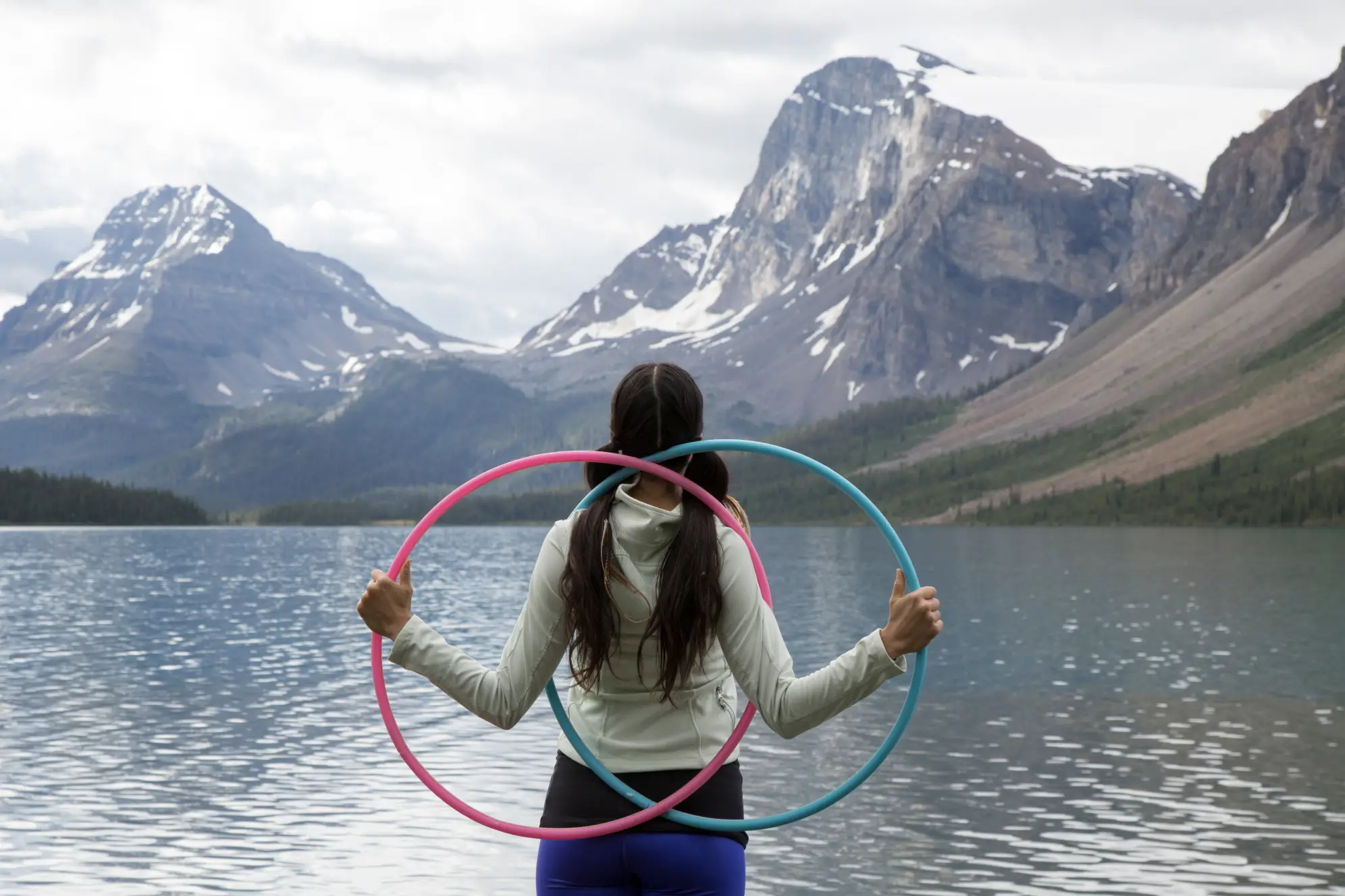 Person with two long pigtails and a blue and pink hoola-hoop standing in front of a lake with mountains in the background