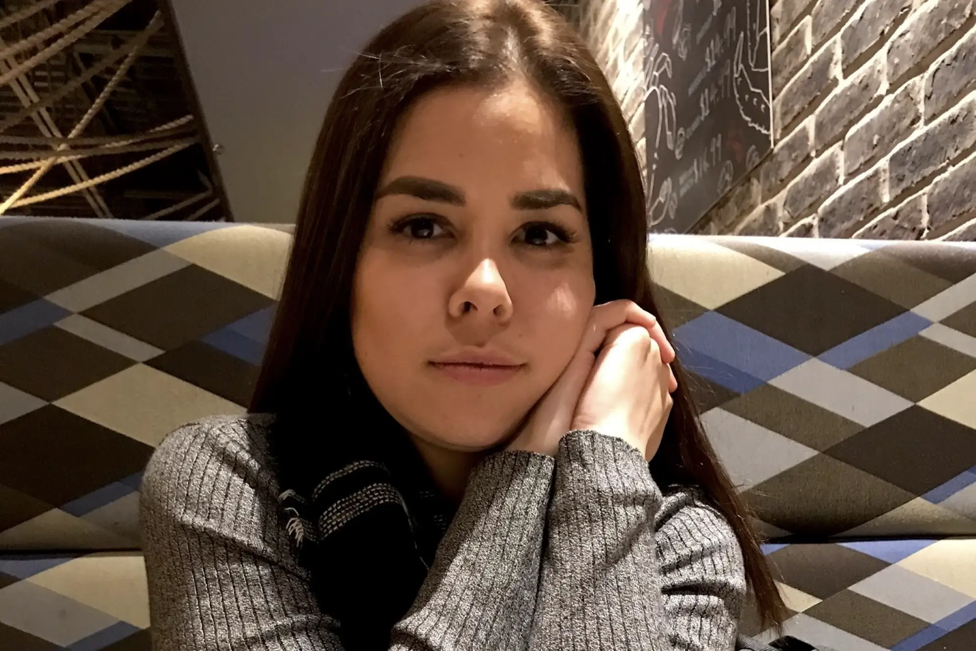 Up-close photo of a young woman looking at camera with a gray sweater sitting on a patterned bench