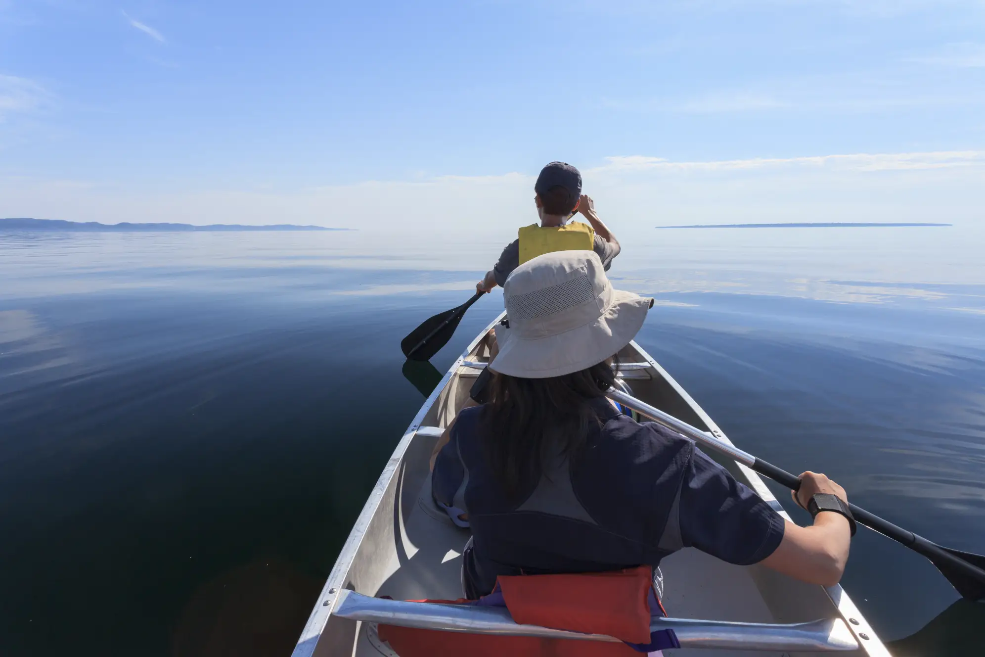Two people paddling in a canoe on a calm lake with a bright blue sky up ahead