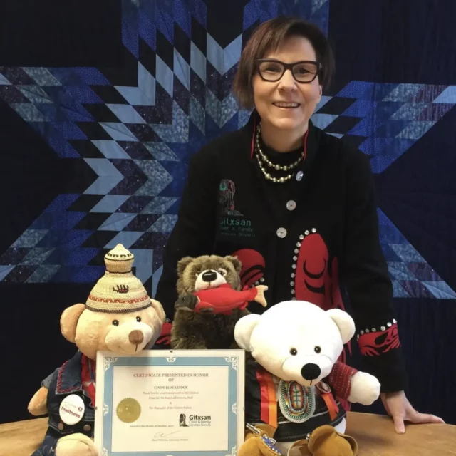 Woman with short brown hair standing in from of a blue quilt with three stuffed animals and certificate on table in front of her