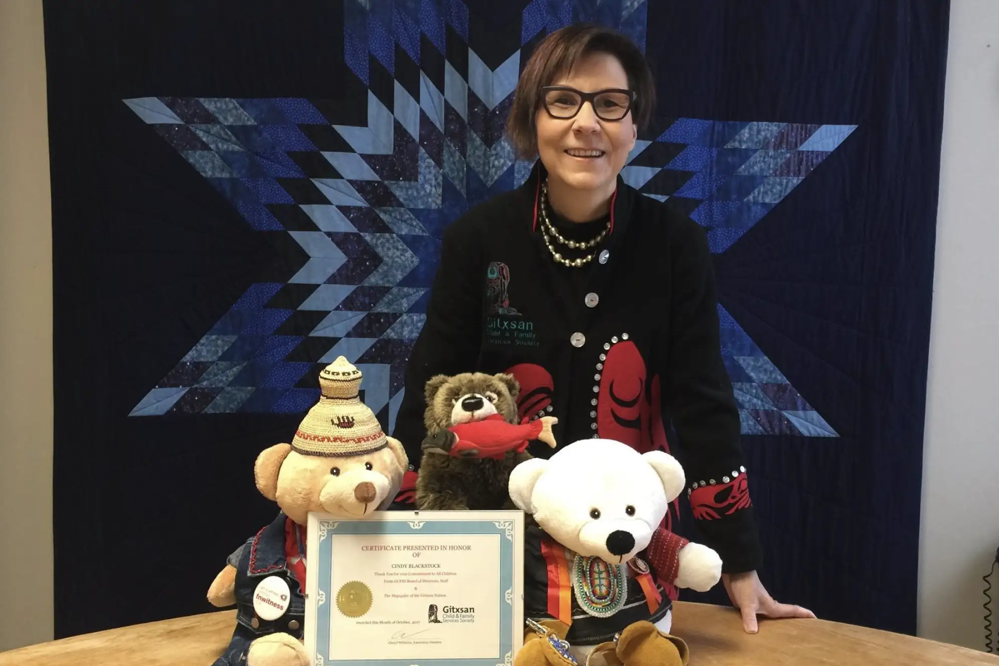 Woman with short brown hair standing in from of a blue quilt with three stuffed animals and certificate on table in front of her