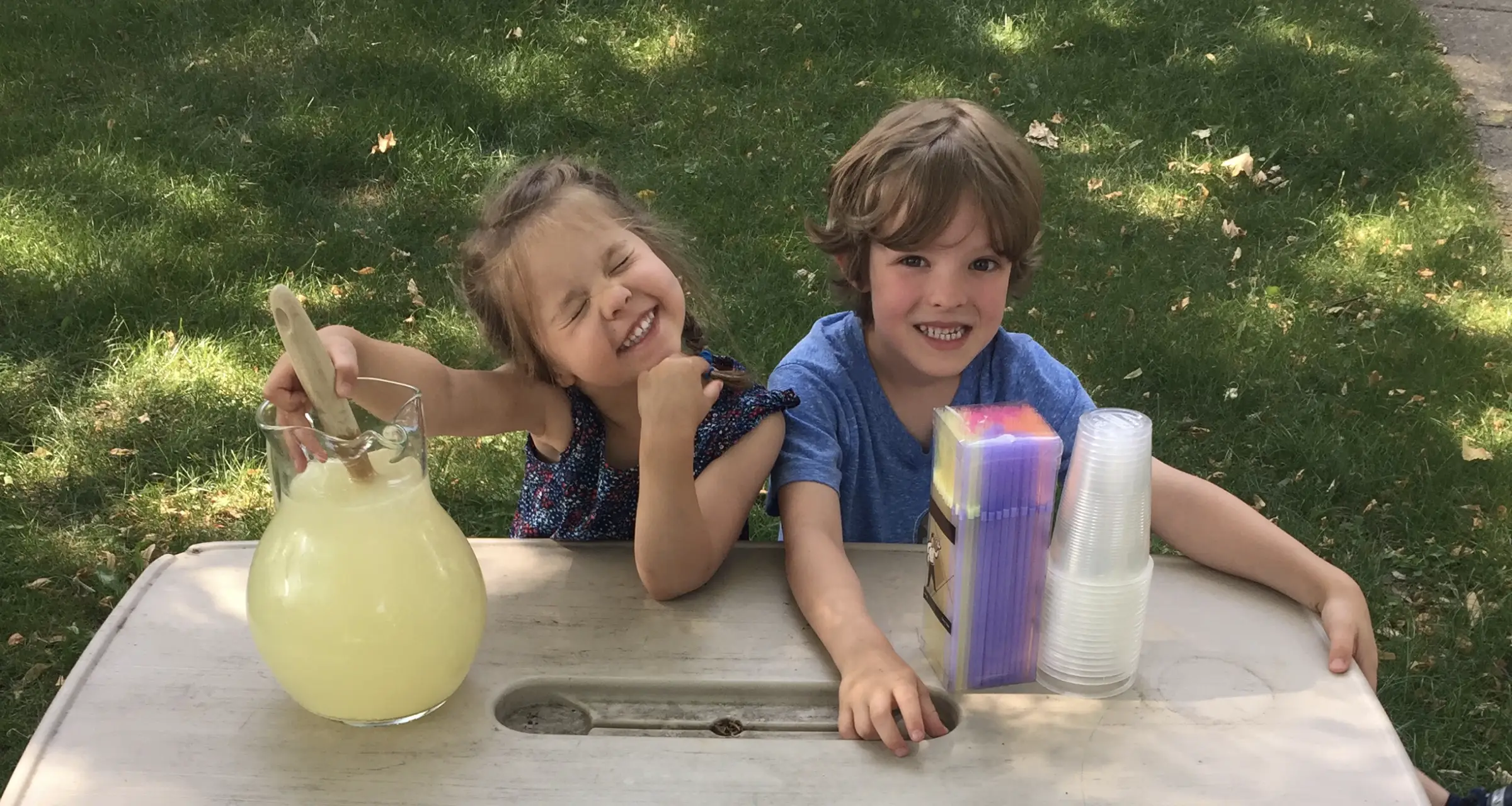 Two children sitting at a table with jug of lemonade and cups and straws