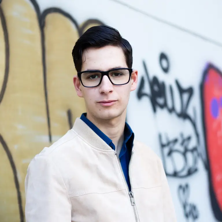 Man wearing glasses standing in front of wall with graffiti and looking at camera