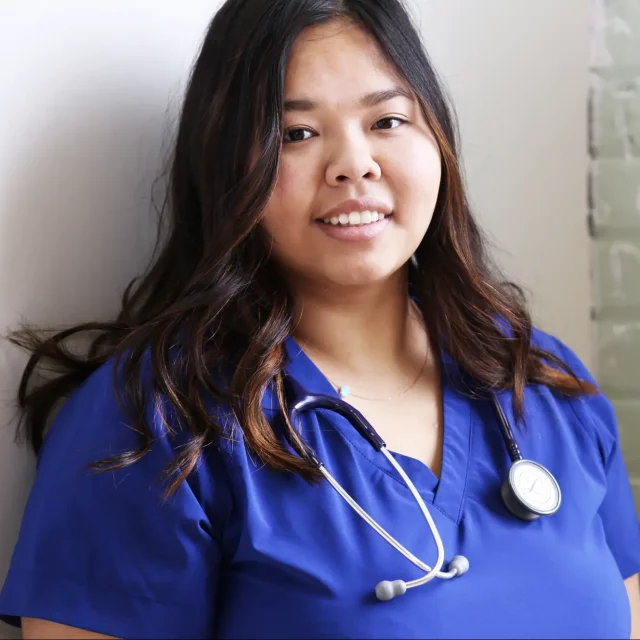 Young woman leaning against white wall wearing dark blue scrubs and smiling at camera