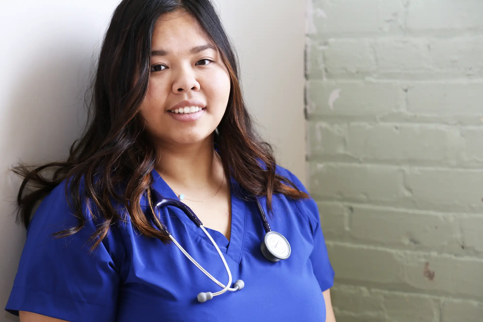 Young woman leaning against white wall wearing dark blue scrubs and smiling at camera