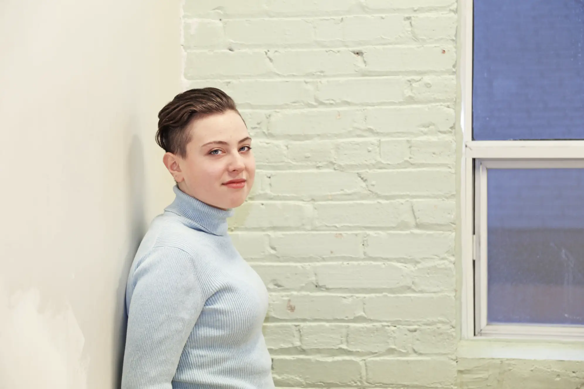 Young woman in grey turtleneck sweater with short hair and smiling at camera while leaning against wall
