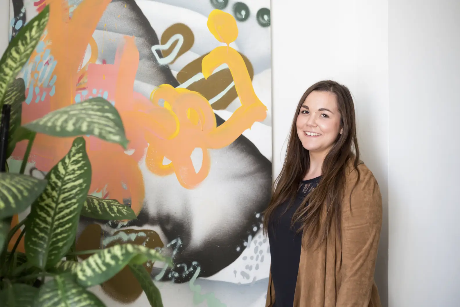 Woman with long hair in sweater smiling and standing in front of painting and beside tall plant