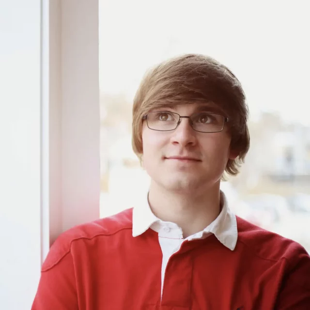 Young man with brown and glasses looking out window and wearing a red shirt