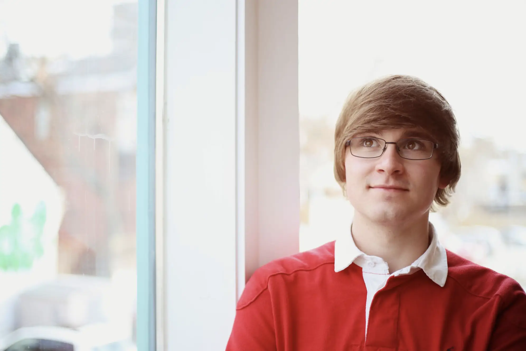 Young man with brown and glasses looking out window and wearing a red shirt