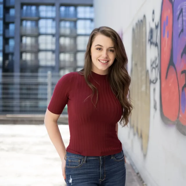 Young woman in burgundy shirt with long hair and standing in front of a wall with graffiti