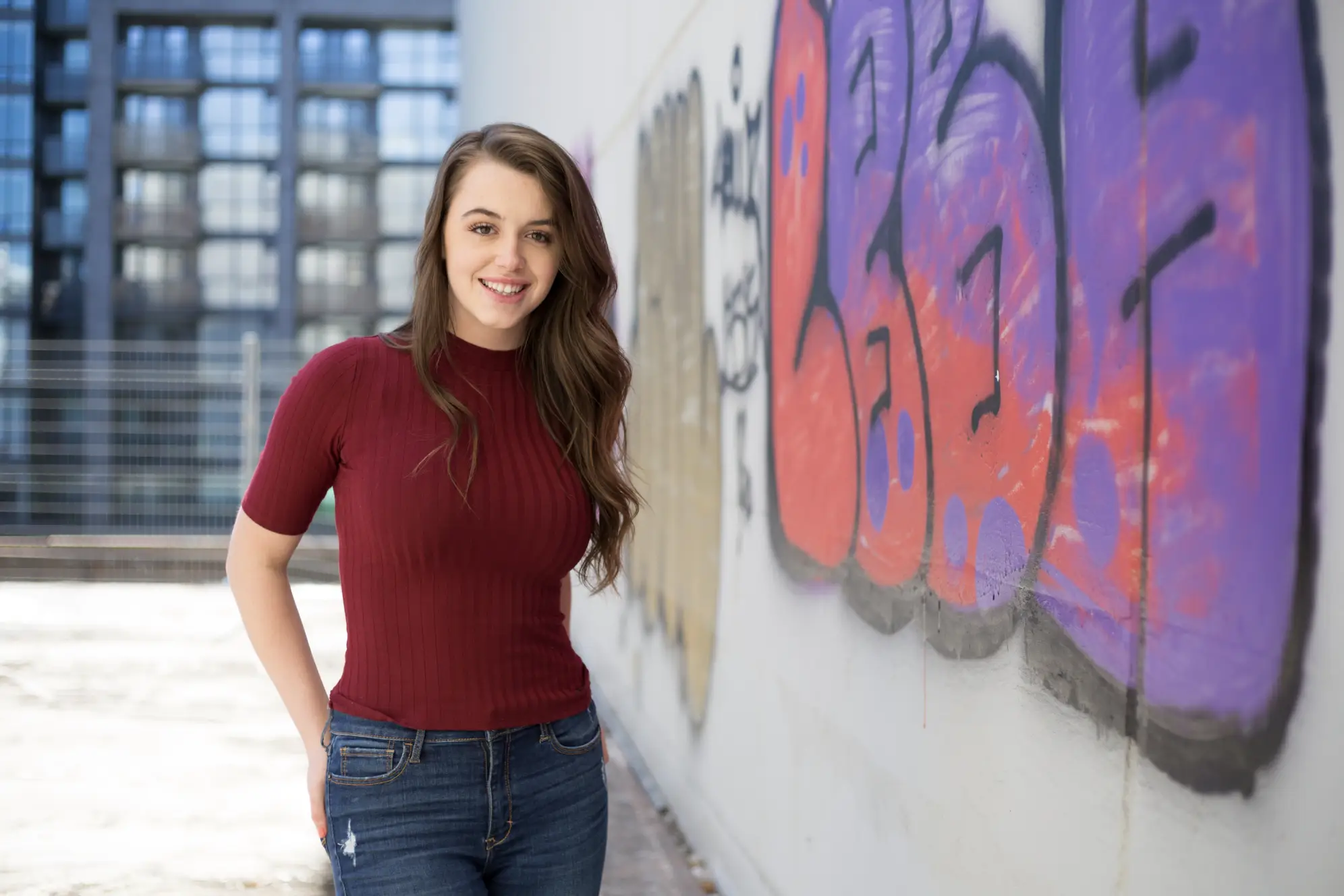 Young woman in burgundy shirt with long hair and standing in front of a wall with graffiti