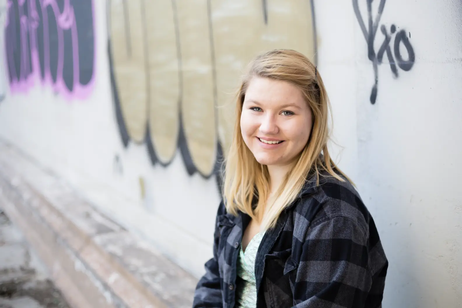 Young woman with long blonde hair sitting in front of wall with graffiti and smiling at camera