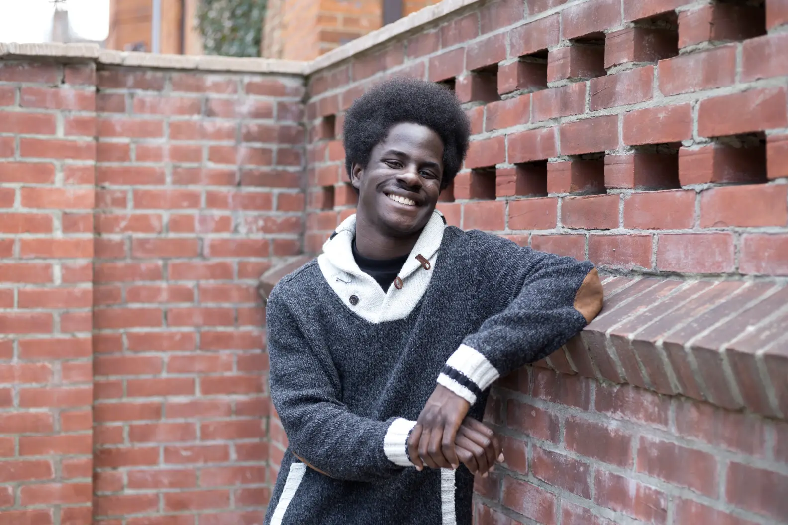 Young man in sweater leaning against a red brick wall and smiling at camera